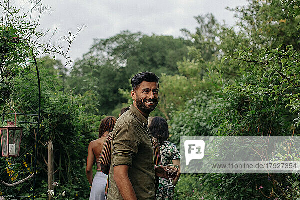 Portrait of happy man holding drink standing amidst plants in garden