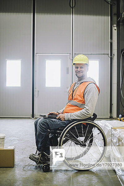 Porträt eines lächelnden Zimmermanns mit Behinderung  der ein digitales Tablet hält  während er im Rollstuhl in einem Lagerhaus sitzt