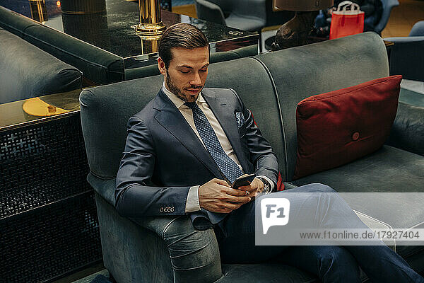 Young businessman text messaging through smart phone while sitting on sofa in hotel lounge