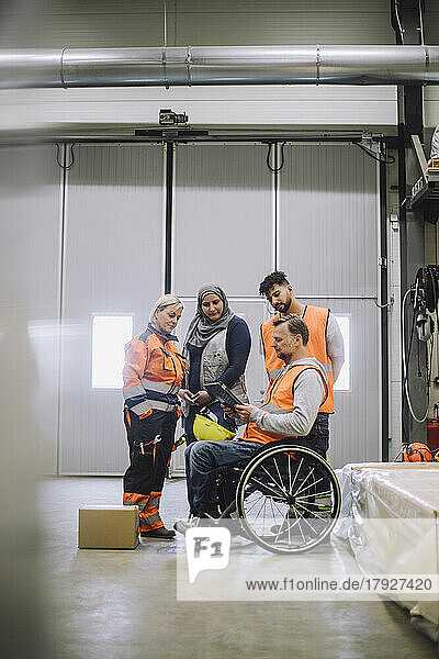 Mature carpenter sitting on wheelchair while discussing over digital tablet with male and female coworkers in warehouse