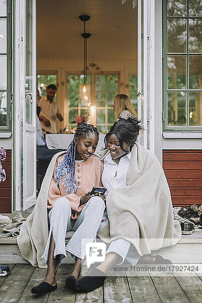 Mutter und Tochter  in eine Decke gehüllt  teilen sich ein Smartphone  während sie auf der Veranda sitzen