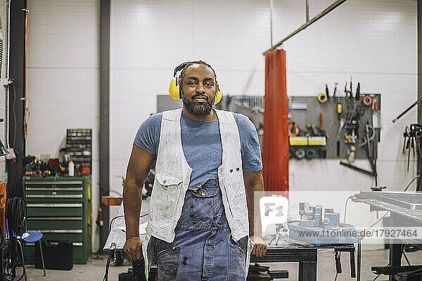Porträt eines lächelnden Tischlers mit Gehörschutz  der sich in einer Werkstatt an eine Werkbank lehnt