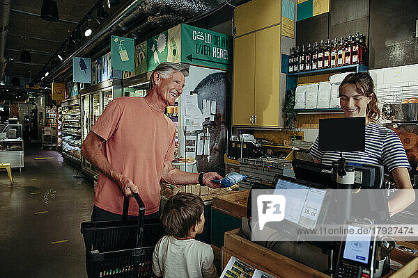 Happy senior man talking to cashier while standing with grandson at checkout counter in supermarket