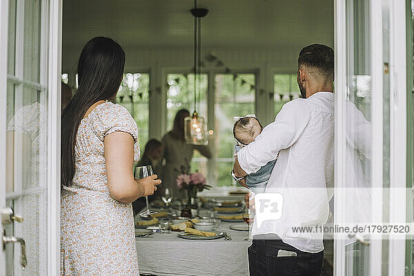 Frau mit Weinglas  die auf einer Party neben einem Mann steht  der seine Tochter trägt  gesehen durch eine Türöffnung