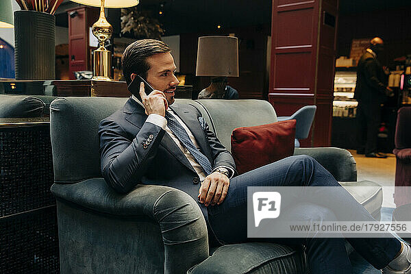 Lächelnder männlicher Berufstätiger  der auf einem Sofa in einer Hotelhalle sitzt und mit einem Mobiltelefon spricht