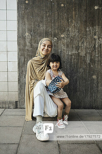 Smiling woman wearing hijab kneeling with daughter in front of wall
