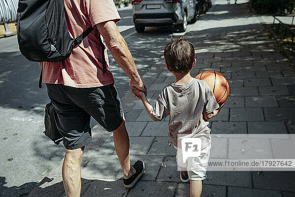 Rückansicht eines Mannes  der die Hand seines Enkels beim Gehen auf dem Bürgersteig hält
