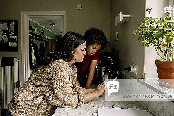 Mutter  die ihr Smartphone mit ihrem Sohn teilt  während sie sich zu Hause auf die Küchentheke stützt