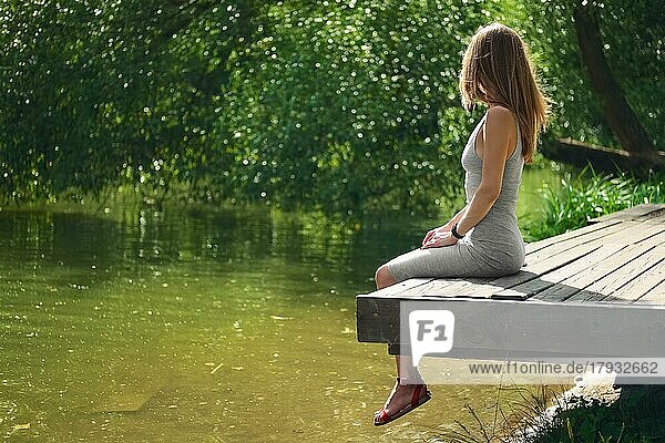 Seitenansicht eines träumenden Mädchens  das auf einem Holzsteg am Flussufer sitzt. Schöne Frau mit vom Winde verwehten Haaren