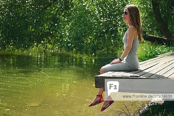 Seitenansicht eines träumenden Mädchens  das auf einem Holzsteg am Flussufer sitzt. Schöne Frau mit vom Winde verwehten Haaren