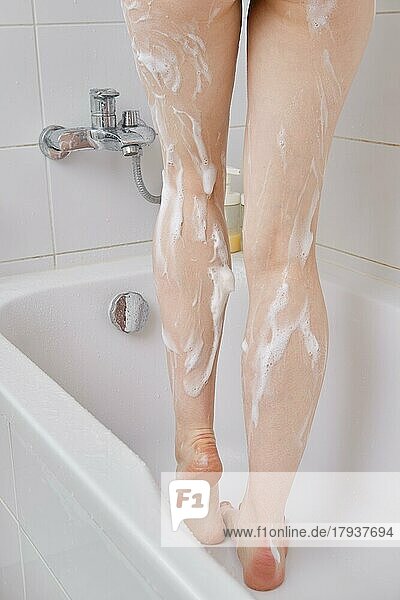 Ausgeschnittenes Foto einer Frau  die Duschgel benutzt  während sie im Badezimmer duscht und in der Badewanne steht. Close up Rückansicht der schlanken weiblichen Beine mit Seifenlauge oder Schaum bedeckt. Hygiene und Körperwäsche Konzept