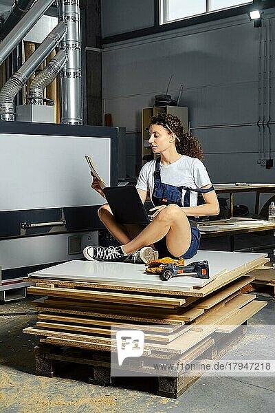Junge Frau in Uniform sitzt auf einem Stapel von Spanplatten mit einem Laptop und einer Papierzeichnung in der Hand