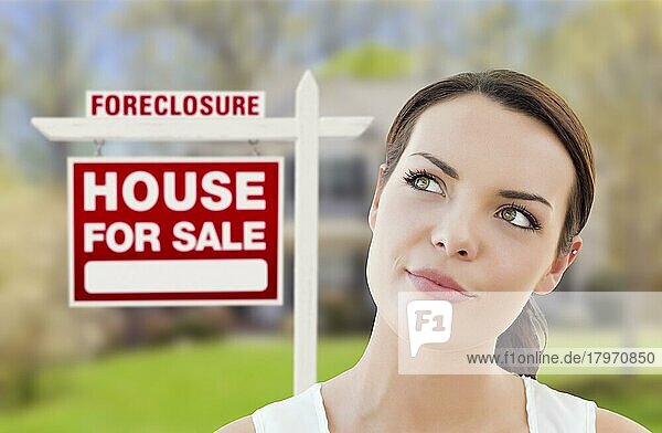 Nachdenkliche hübsche gemischtrassige Frau vor Haus und Zwangsversteigerung Haus zu verkaufen Immobilien Schild nach oben und zur Seite schauen