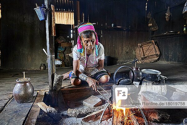 Kayan-Lahwi-Frau mit Messinghalswendeln und traditioneller Kleidung  die in ihrem Haus Wasser zum Kochen bringt  um ihrem Lieblingsgetränk  einem Likör aus vergorenem Sorghum (links im Glas)  Pan Pet Region  Kayah State  Myanmar  Asien