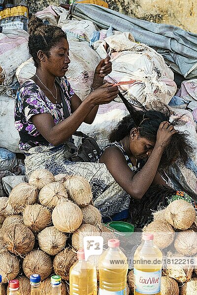 Two woman braiding hair  market stall  coconuts  bags of charcoal behind  Mahajanga  Madagascar  Africa