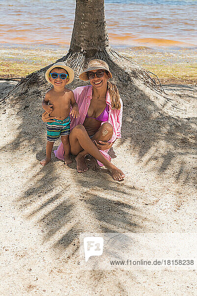 Mutter und kleiner Junge posieren unter einer Palme am Strand mit Sonnenbrille