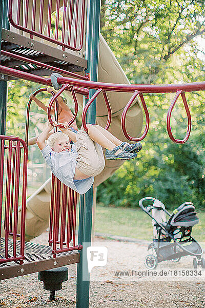 5 Jahre alter weißer  blonder Junge hängt auf einem Spielplatz an einem Klettergerüst.