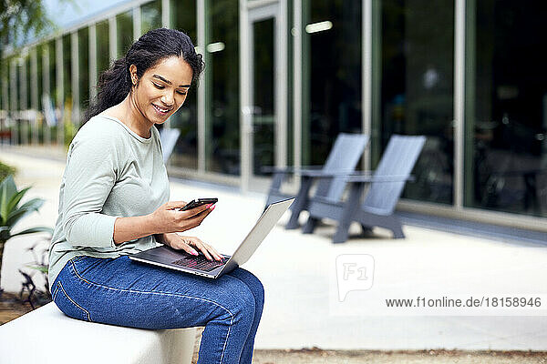 Lächelnde Geschäftsfrau  die ein Smartphone benutzt  während sie mit einem Laptop sitzt