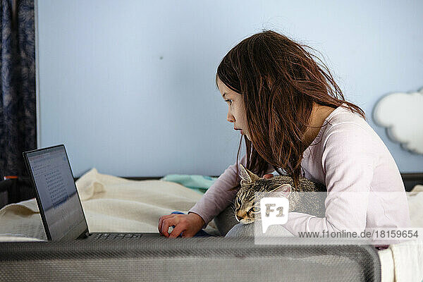 A little girl sits on bed with cat in lap looking at a computer