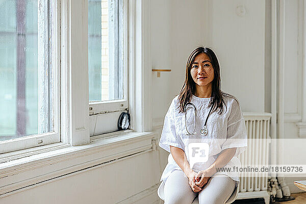 Porträt einer selbstbewussten Ärztin  die in einer Klinik am Fenster sitzt