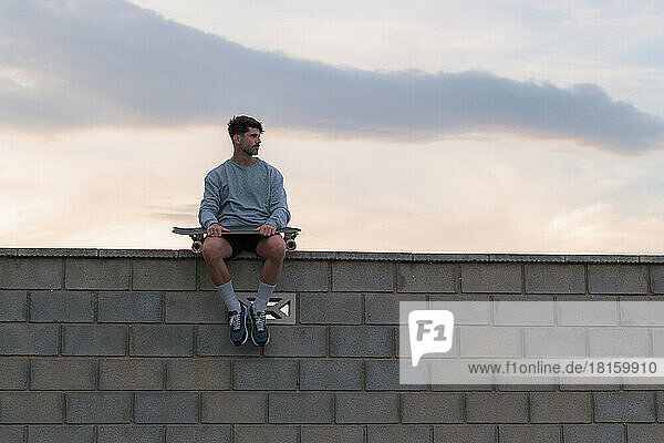 Junger Mann sitzt bei Sonnenuntergang mit einem Skateboard auf einer Mauer