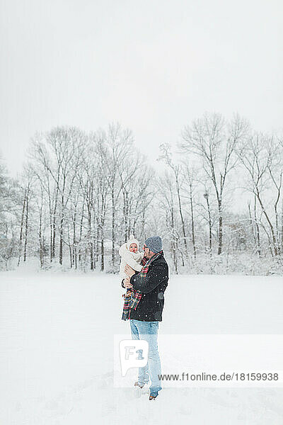 30-year-old father holds up 12-month-old baby in snowy field.