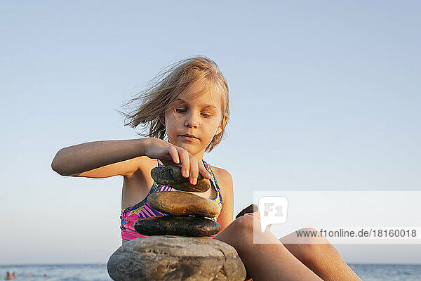 Ein Mädchen sitzt bei Sonnenuntergang am Meeresufer und spielt mit Steinen.