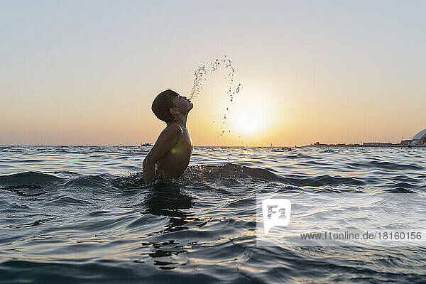 Ein Junge springt bei Sonnenuntergang ins Meer und spritzt sich das Wasser aus dem Mund.