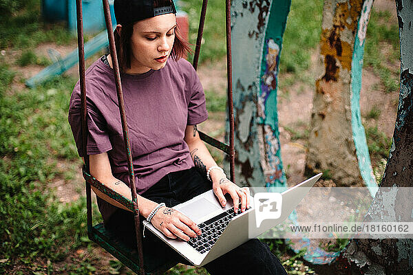 Junge Studentin mit Laptop auf einer Schaukel sitzend im Sommer