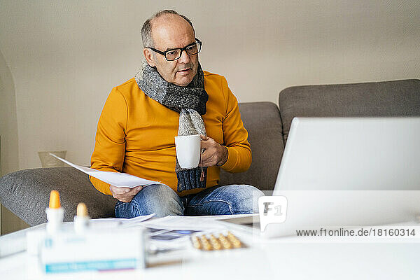Senior man with mug and medical report looking at laptop in living room