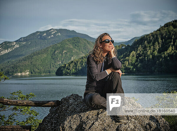 Smiling thoughtful woman sitting on rock in front of lake
