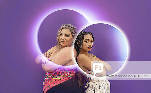 Women standing with arms crossed seen through glowing circles in front of purple wall
