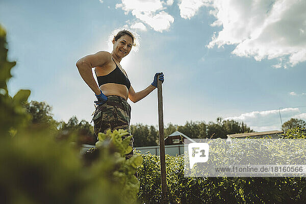 Smiling mature woman holding shovel standing with hand on hip in vegetable garden