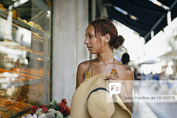 Hungrige junge Frau mit Hut blickt durch das Schaufenster eines Feinkostladens