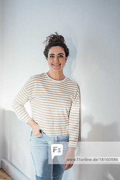 Smiling woman with hand in pocket leaning on white wall at home