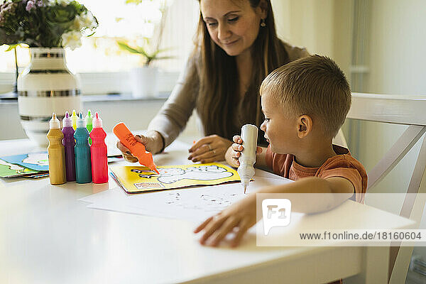 Mother coloring with paint tube in book by son at home