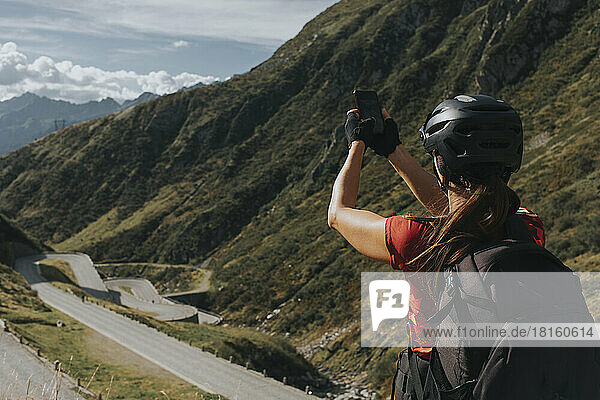 Woman wearing helmet photographing Gotthard Pass through smart phone on sunny day