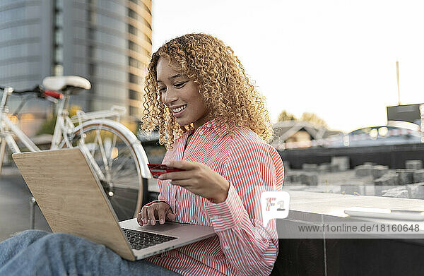 Smiling woman with credit card doing online shopping on laptop by bench