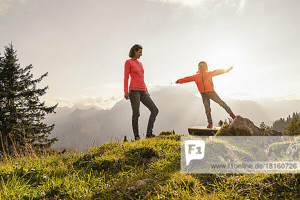 Smiling mother looking at daughter standing with arms outstretched on bench
