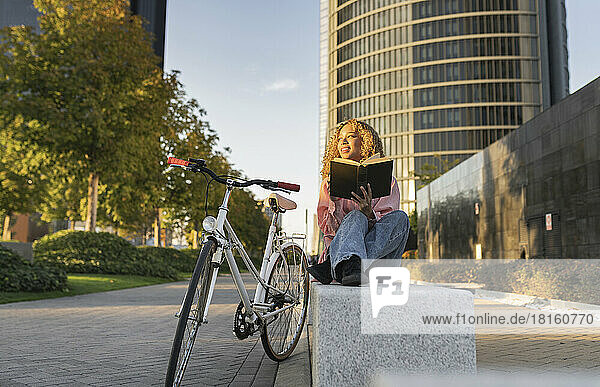 Smiling thoughtful woman with book sitting on bench by bicycle
