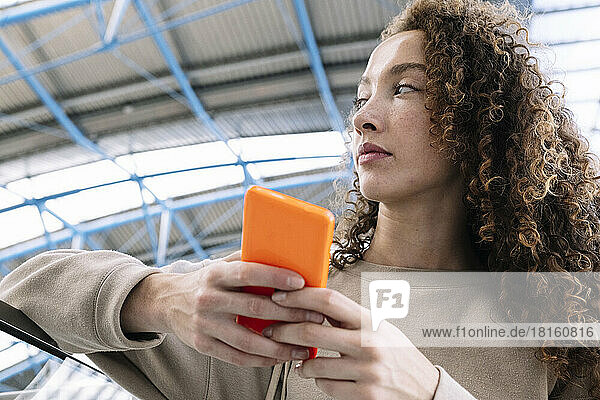 Nachdenkliche Frau mit lockigem Haar hält ihr Mobiltelefon am Geländer in der Hand