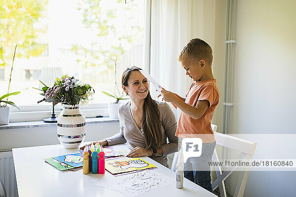 Smiling mother looking at son holding paper standing on chair