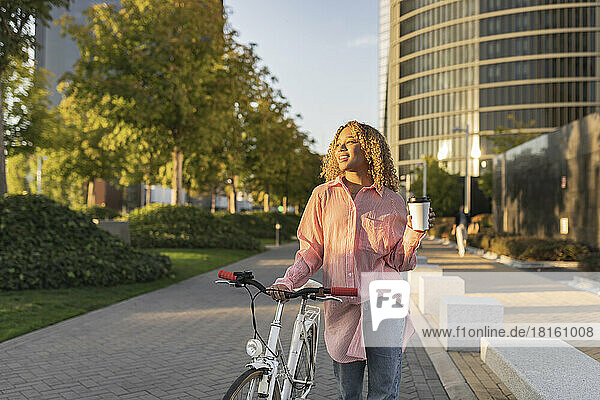 Lächelnde Frau hält Einwegkaffeetasse in der Hand und läuft mit dem Fahrrad auf dem Fußweg