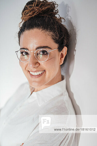 Smiling woman with eyeglasses leaning on white wall at home