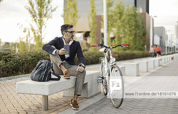 Businessman with smart phone sitting on bench by bicycle