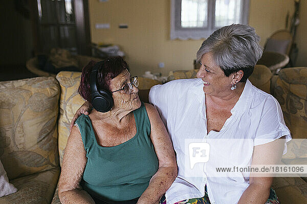 Lächelnde Frau blickt Mutter mit Kopfhörern an  die zu Hause auf dem Sofa sitzt