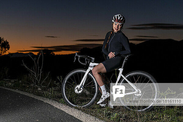 Lächelnder Radfahrer stützt sich in der Abenddämmerung auf sein Fahrrad in der Nähe der Straße
