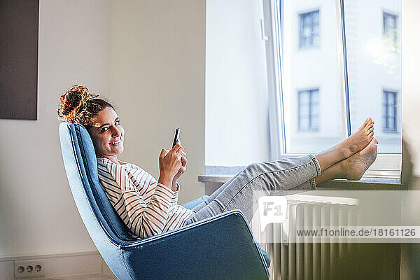 Smiling woman holding mobile phone with legs on window sill home