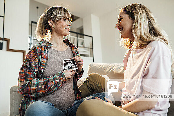 Smiling sister talking to pregnant woman holding ultrasound at home