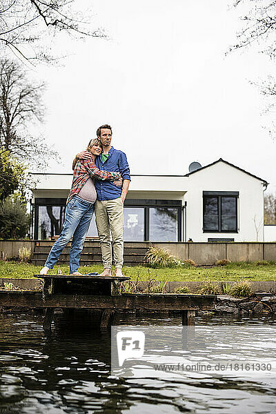 Expectant couple standing together on jetty at lake in front of house